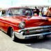 A vintage red 1957 Chevrolet showcased at an outdoor car exhibition with attendees.